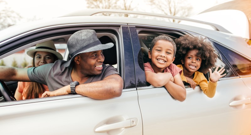 A family of four in a car with the father and 2 smiling kids in the back peering out the window